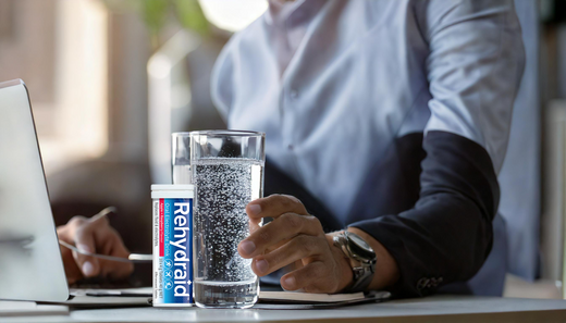 A professional working at a desk, with a glass of water and a dissolving Rehydraid effervescent tablet, representing hydration and mental clarity.